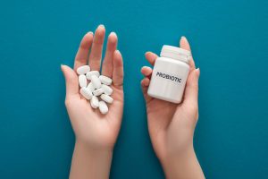 Cropped view of woman holding white probiotic container and pills in hands on blue background.
